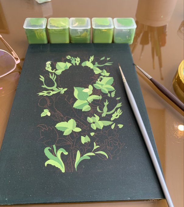 An in-progress painting on black paper featuring green floral patterns, with small containers of green watercolor paint and two brushes visible on the workspace.