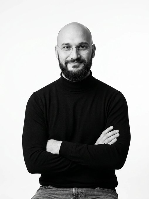 A black and white portrait of a bald man with a beard and glasses, wearing a black turtleneck sweater, posing with his arms crossed against a plain white background.