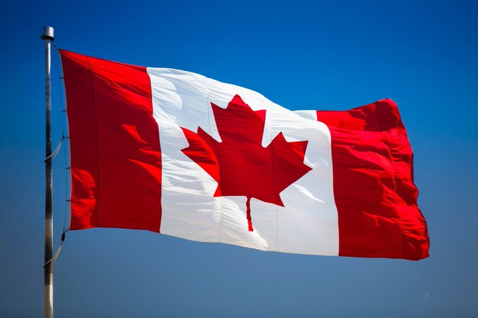 The national flag of Canada featuring its iconic red maple leaf on a white band between two red stripes, waving against a clear blue sky.