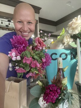 A smiling bald woman in a blue top holds a large bouquet of pink and white flowers in a kitchen setting.