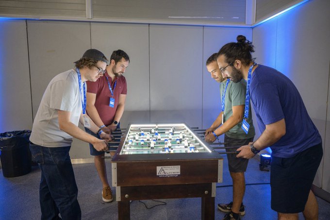 Four men wearing lanyards are gathered around and playing a brightly lit foosball table.