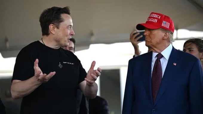 Elon Musk (wearing a black "SpaceX" t-shirt) speaks with animated hand gestures while Donald Trump, wearing a navy suit, red tie, and a red "AMERICA AGAIN" cap with an American flag, listens with a serious expression.