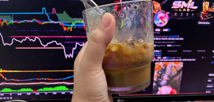 A hand holds a glass of iced coffee in front of a monitor displaying colorful cryptocurrency trading charts and an 'SML LAB' interface.