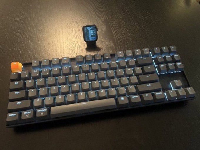 A custom mechanical keyboard with dark keycaps, RGB backlighting, and a small OLED display module, resting on a dark wooden desk.