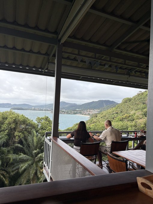 An open-air seaside restaurant terrace overlooking a bay and green hills, with diners seated at tables under a corrugated roof.
