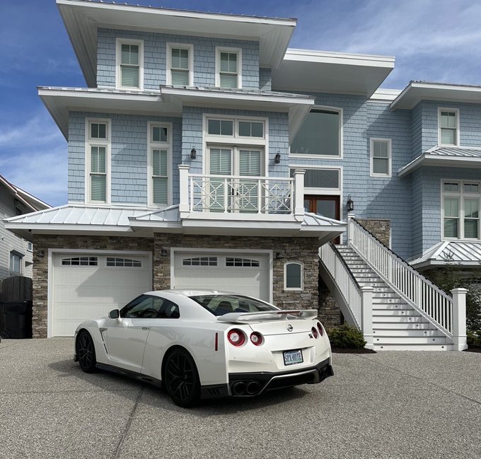 A white Nissan GT-R sports car is parked on a paved driveway in front of a large, light blue house with multiple levels, a balcony, and a two-car garage.