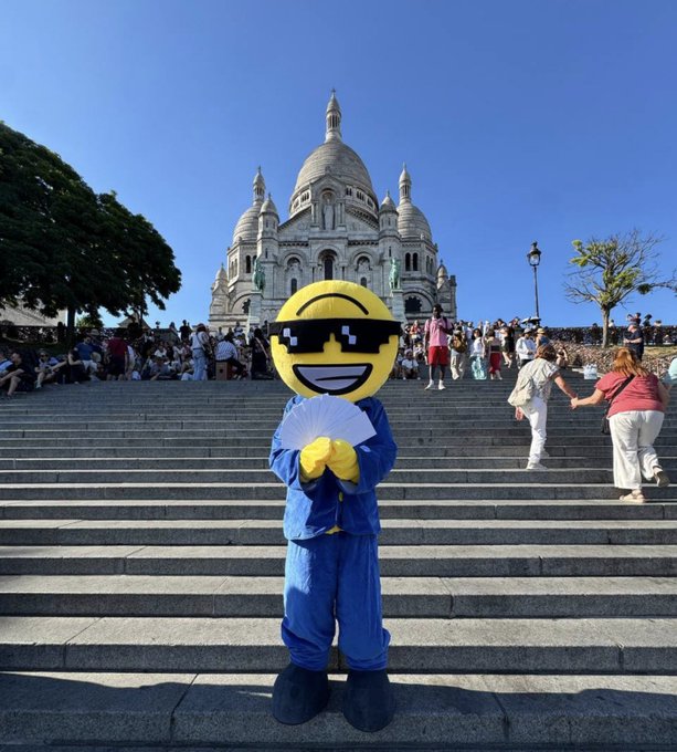 A person in a yellow emoji "cool" sunglasses mascot costume with a folding fan stands on the steps of Sacré-Cœur Basilica in Montmartre, Paris, promoting a memecoin meetup and a treasure hunt for 15 hidden envelopes containing €50 each.