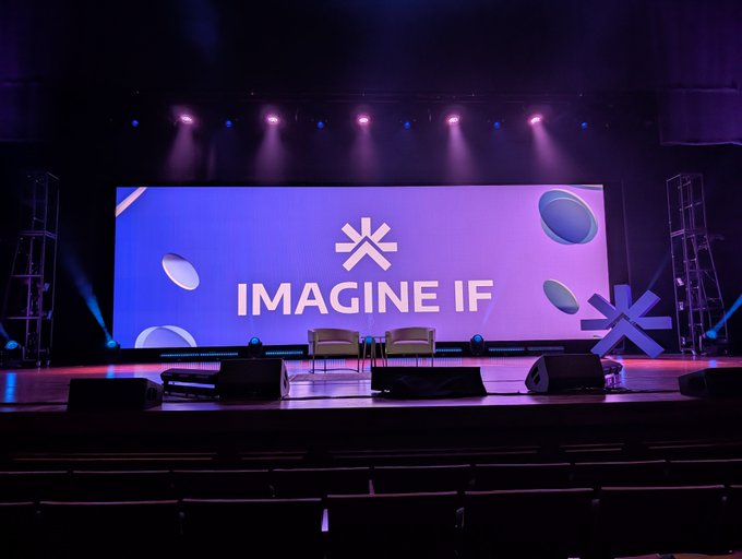 An empty auditorium stage set up for an event, with a large purple-lit screen displaying 'IMAGINE IF' and an asterisk-like logo, illuminated by stage lighting.