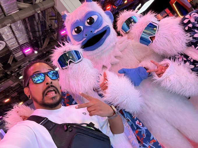 A man wearing reflective blue sunglasses poses with the white furry "Lofi the Yeti" mascot, who is also sporting matching blue ski goggles, at a lively Token2049 event filled with colorful booths and lights.