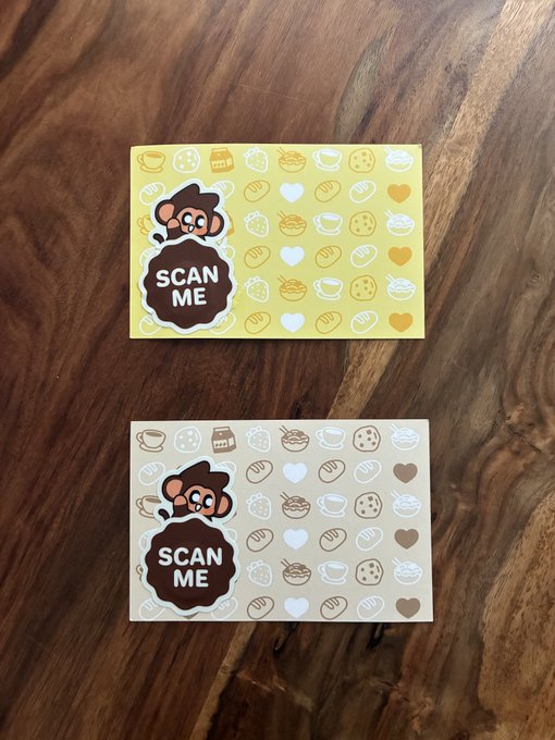 Two loyalty or business cards featuring a monkey mascot peeking out from a 'SCAN ME' speech bubble, placed on a wooden surface; the top card is yellow and the bottom is a brown/kraft tone.