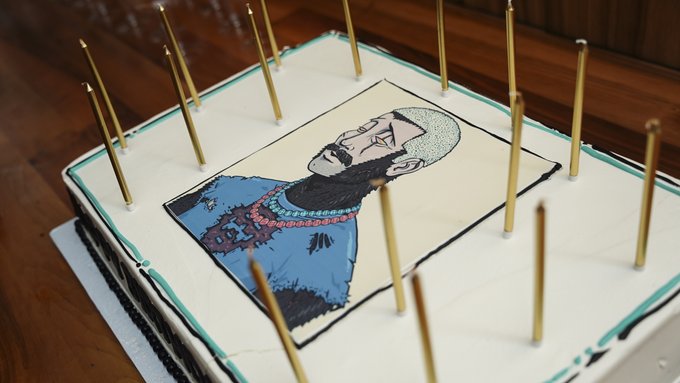 A decorated birthday cake featuring an edible image of a bearded man wearing sunglasses and a rosary, topped with tall, thin candles.