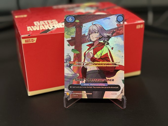 A trading card titled "Shoo's Perseverance" featuring an anime-style character, displayed on a clear stand in front of a red "GATES AWAKENING" booster box with an "AGES 13+" label.