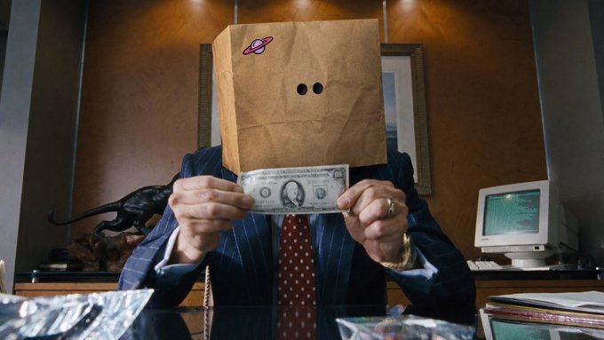 A person in a pinstripe suit with a red tie sits at a desk holding a one-dollar bill in front of their face, which is concealed by a brown paper bag decorated with a small planet sticker and two eye holes. The image thematically represents the post's call for alpha team members who deliver genuine, well-researched calls rather than superficial "easy 3x" suggestions—symbolizing the value of real substance (the dollar) behind the hidden identity, much like the credible DD being sought over empty hype.