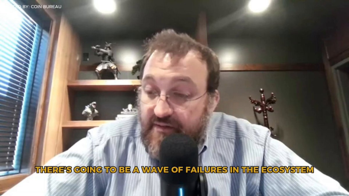 The image shows a man wearing glasses sitting at a desk with a microphone, likely preparing for an interview or participating in a live webinar. The background features a window that allows natural light into the room. There are also several figurines on display behind the man, adding a personal touch to the scene.