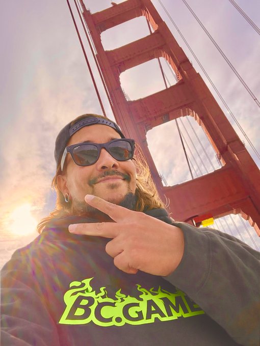 A man wearing sunglasses and a 'BC.GAME' hoodie makes a peace sign in front of the Golden Gate Bridge at sunset.
