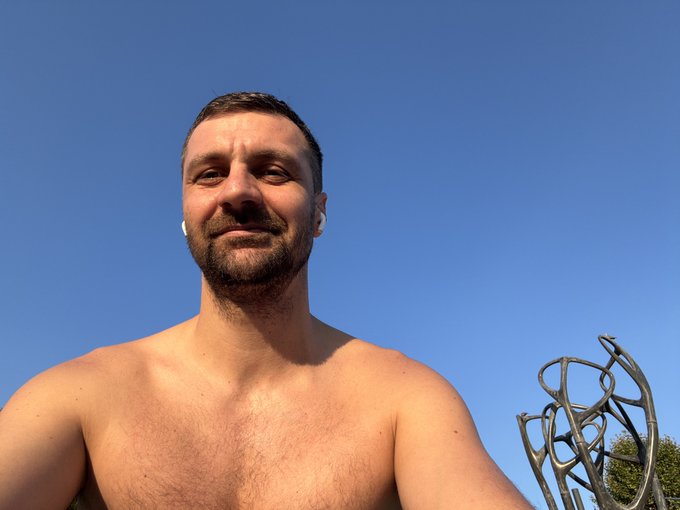 A low-angle selfie of a shirtless, bearded man outdoors under a clear blue sky, wearing white wireless earbuds, with a metal abstract sculpture visible in the background.