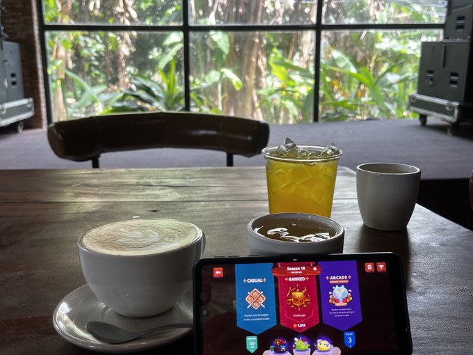 A coffee shop scene with a cappuccino, iced tea, and a small ceramic cup on a wooden table, with a tablet displaying a card game showing 'CASUAL', 'RANKED', and 'ARCADE' modes against a backdrop of tropical greenery through large windows.