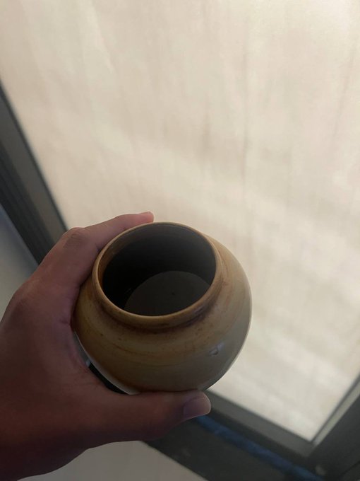 A hand holding a small ceramic pot or vessel with a tan glaze, viewed from above against a light-colored background.
