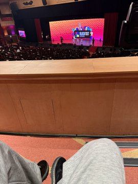 A seated attendee’s legs face a large conference auditorium with a bright red stage screen and rows of audience members.