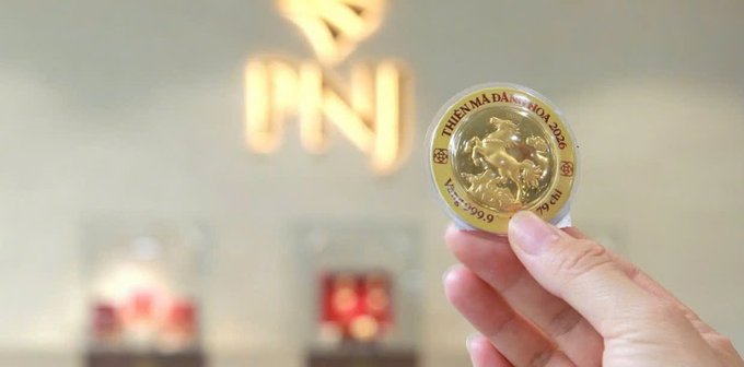 A hand holds up a gold commemorative coin featuring a bull design and inscribed 'THIÊN MÃ ĐÁI KHOA 2020' and '9999.99' in front of a blurred background.