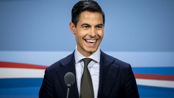 A man in a navy suit and patterned tie smiles broadly at a podium with a microphone, set against a blue backdrop with red and white stripes. The caption relates to the post's alarm about potential new taxes on unrealized gains, suggesting the smiling politician may be delivering or celebrating such a policy announcement.