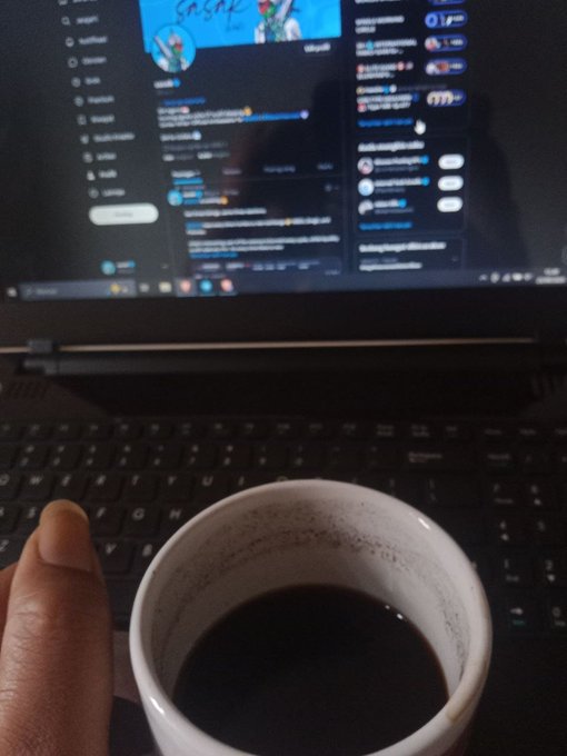 A first-person view of someone holding a white mug of black coffee while sitting in front of an open laptop displaying a social media feed.