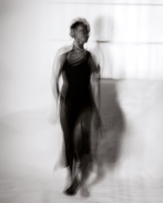 A black-and-white motion-blurred photograph shows a woman in a dark dress with a flowing sheer fabric, creating a ghostly, ethereal effect.
