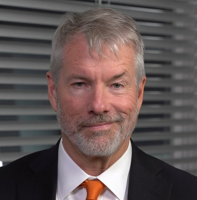 A professional headshot of an older man with gray hair and a beard, wearing a dark suit, white shirt, and orange tie, photographed in front of horizontal window blinds.