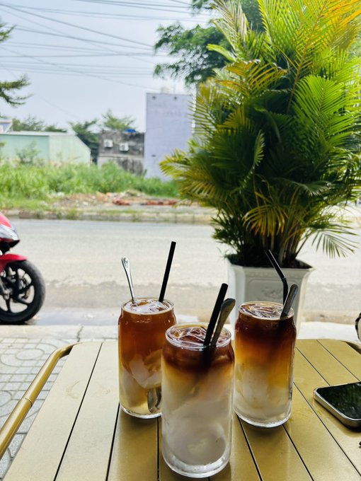 Three glasses of iced coffee with black straws and spoons sit on a yellow outdoor table, with a potted palm plant, a motorbike, and a street visible in the background.