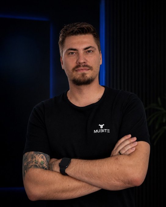 Peter Andreas, founder of Mubite, stands confidently with arms crossed wearing a black "MUBITE" branded t-shirt and smartwatch, complementing the post's introduction of him as the visionary driving the prop firm. The portrait's professional, tech-forward aesthetic with moody blue lighting reinforces the post's theme of building credibility and infrastructure in crypto trading.