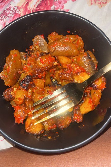 A black bowl filled with a spicy dish of meat or potato chunks coated in red chili paste and seeds, with a metal fork resting inside.