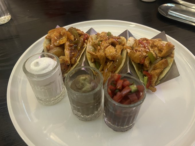 A white plate on a dark wooden table displaying three chicken fajita tacos in metal holders, accompanied by small glasses of sour cream, guacamole, and salsa.