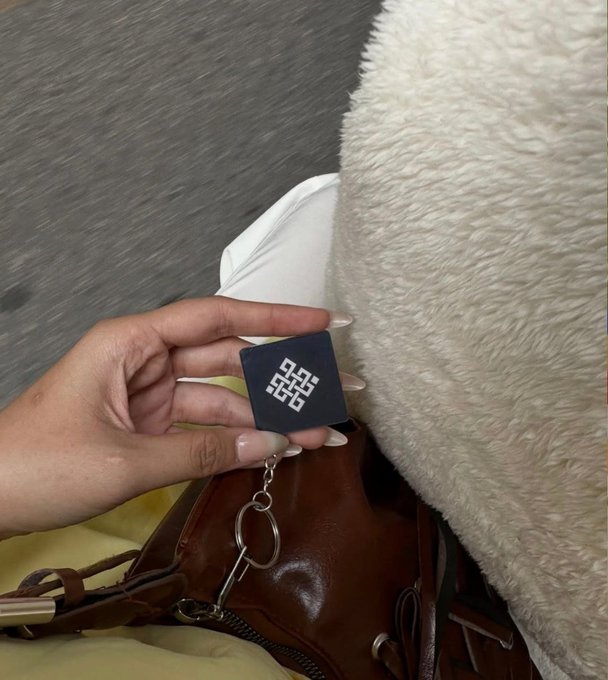 A hand holding a black QR code keychain over a brown leather bag and white fluffy blanket.