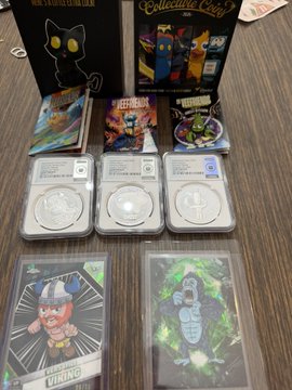 A collection of graded silver coins displayed in protective slabs (graded 70 by what appears to be PCGS/NGC) arranged on a wooden surface, accompanied by various trading cards and collector's boxes including a Marvel Collector Corps box.