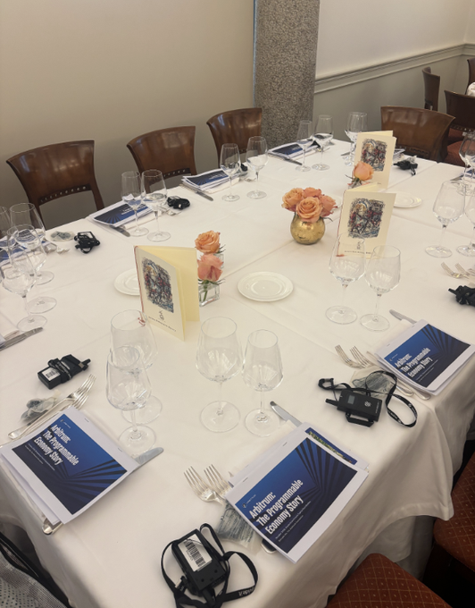 A formally set dining table features placards reading "Arbitrum: The programmable company theory," with wine glasses, cameras, and floral centerpieces arranged in preparation for an Arbitrum investor event attended by finance leaders and banks.