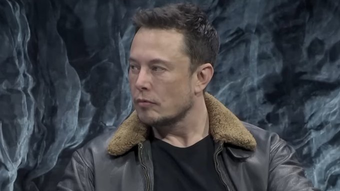 Elon Musk wearing a brown leather jacket with a shearling collar, looking to the side against a dark textured background.