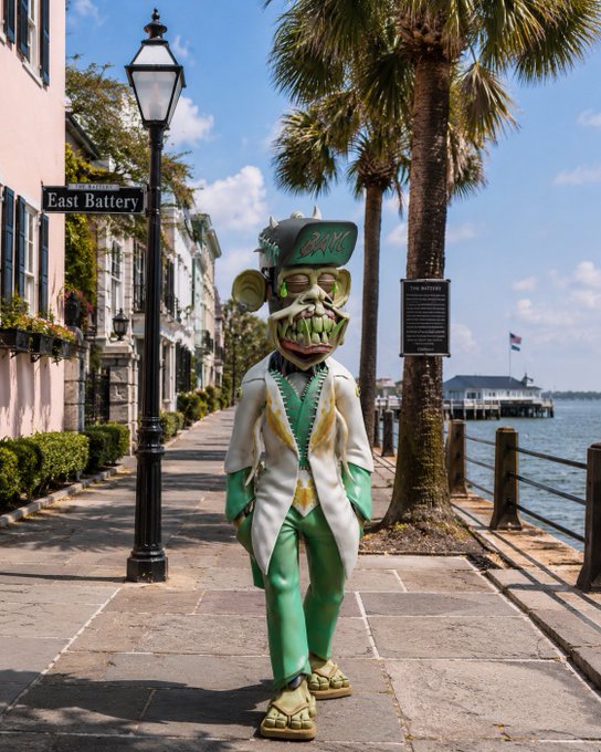 A person in an elaborate green and white suit with a grotesque carved mask standing on East Battery street in Charleston, with palm trees and a waterfront pier in the background.