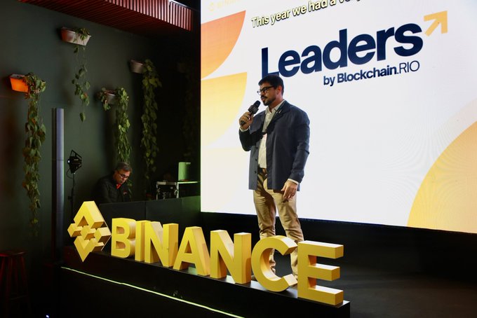 A man in a blazer speaks into a microphone on a stage displaying the text "Leaders by Blockchain.RIO", with a large illuminated "BINANCE" sign in the foreground.