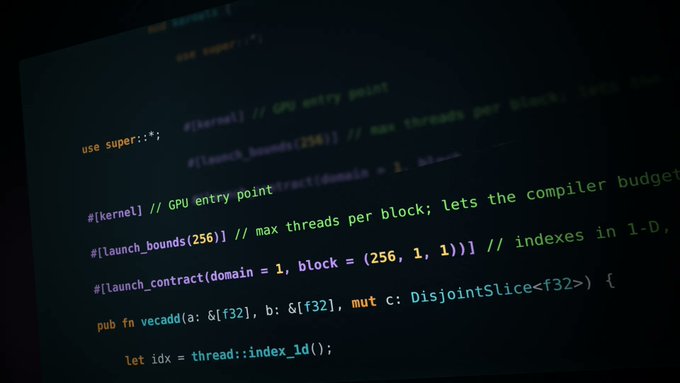 A close-up photo of a computer monitor displaying Rust code with GPU kernel annotations like "#[kernel]", "#[launch_bounds(256)]", and "#[launch_contract(...)]".