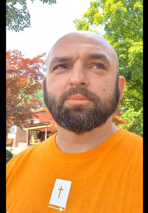 A close-up selfie of a bald man with a beard wearing an orange t-shirt featuring a small book-and-cross graphic, standing outdoors with green trees and a red-leaf tree behind him.