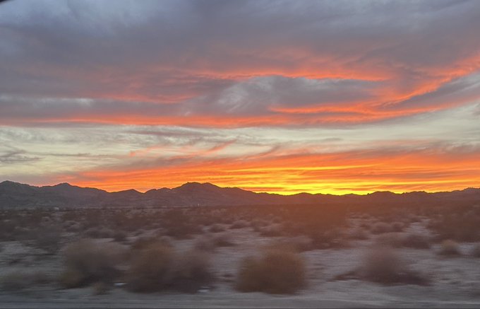 A stunning desert sunset captures vibrant orange and red clouds streaking across the sky over a silhouetted mountain range and arid landscape with desert shrubs.
