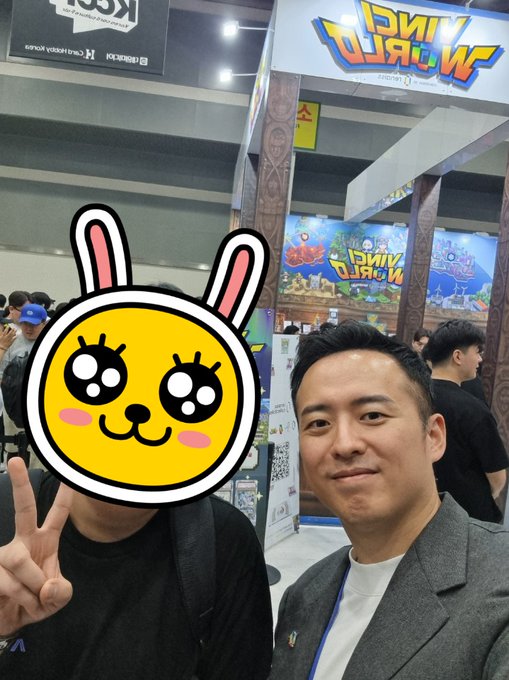 The image features two men posing for a picture with cartoon bunny faces as their masks, which are part of a larger promotional event at a convention center. The background includes a booth named " Renaiss @Plus_Ultra_715" and another person standing in front of it. One man is wearing a suit jacket while the other has a backpack on his back.