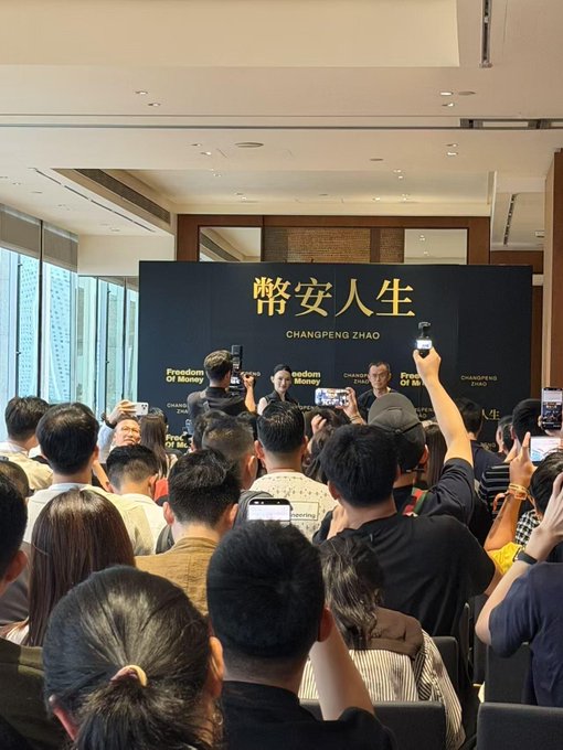 A crowd of people hold up phones to film speakers at a podium bearing the text "常安人生 CHANGPENG ZHAO Freedom Of Money."