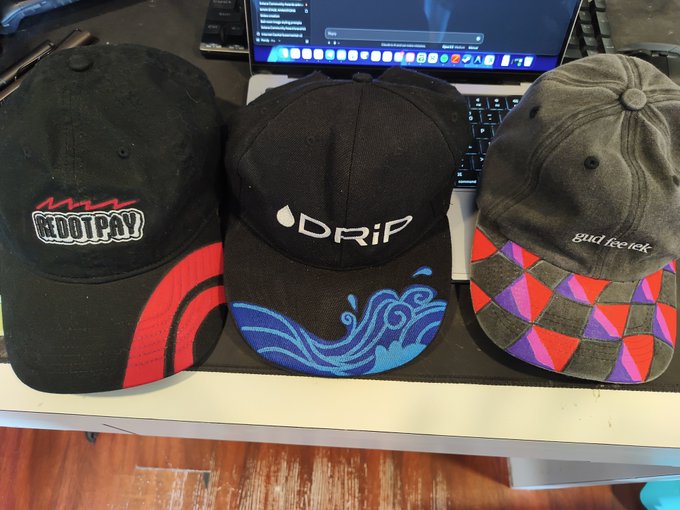 Three baseball caps displayed on a white desk in front of a laptop: a black cap with 'BOOPAY' text, a black cap with 'DRiP' and blue wave embroidery, and a dark green cap with a purple and red checkered brim and 'gud feed' embroidery.