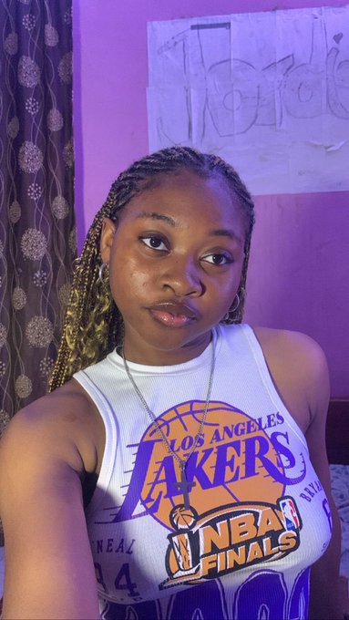 A young woman with braided hair wearing a white 'LOS ANGELES LAKERS NBA FINALS' tank top, with 'Thank God' written on paper behind her.