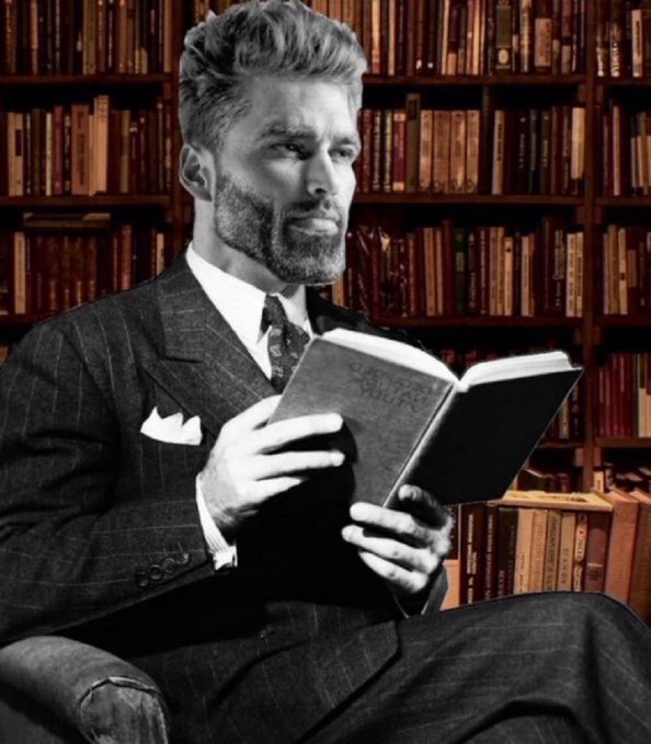 A black-and-white photo of a well-dressed bearded man in a pinstripe suit reading a book in front of a large bookshelf.