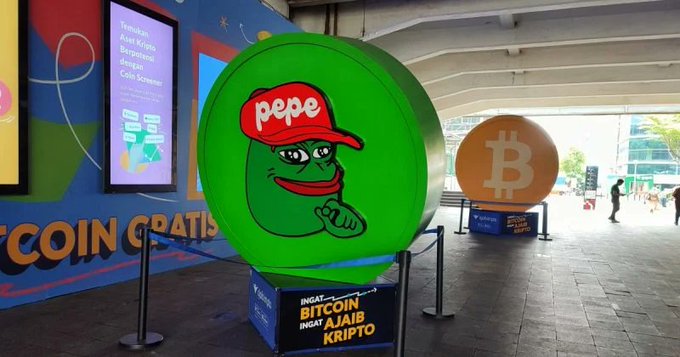 A display featuring a large inflatable green Pepe the Frog coin character with a red 'pepe' cap, alongside a Bitcoin coin sculpture, with a sign reading 'INSERT: BITCOIN / AJAIB / KRIPTO' and an advertisement mentioning 'Coin Scanner' in Indonesian.