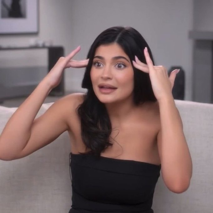 Kylie Jenner sits on a beige couch wearing a black strapless top, gesturing with both hands near her head while speaking.