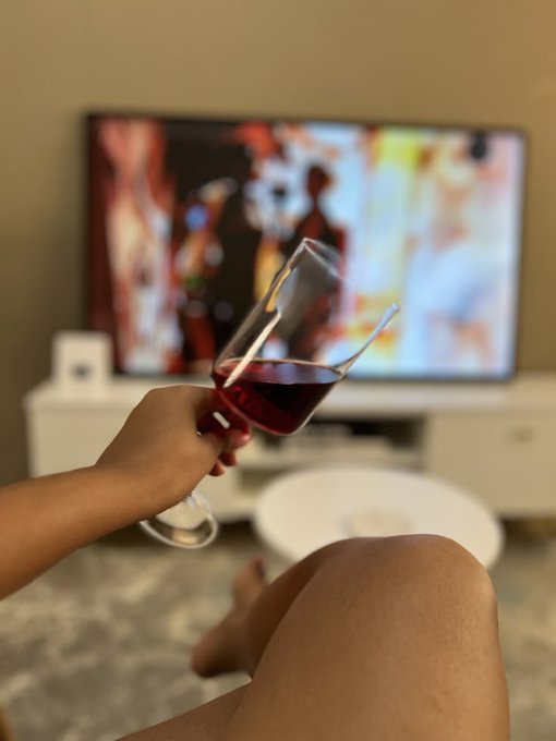 A person holds a glass of red wine while watching a blurred television, with their legs visible in the foreground in a cozy living room setting.