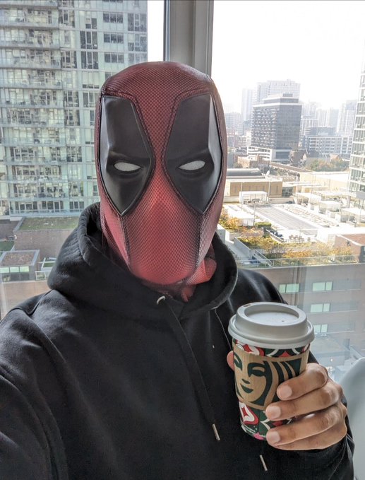 A person wearing a Deadpool mask and black hoodie holding a Starbucks coffee cup while taking a selfie in front of a city skyline visible through a window.
